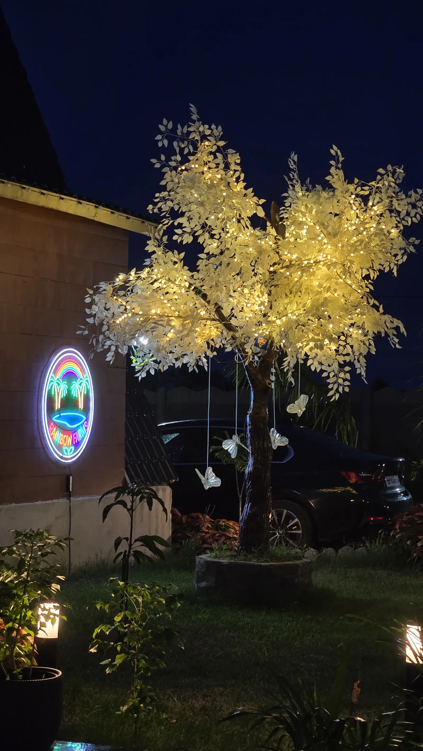 A glowing white tree beside the neon Rainbow Farms sign at dusk