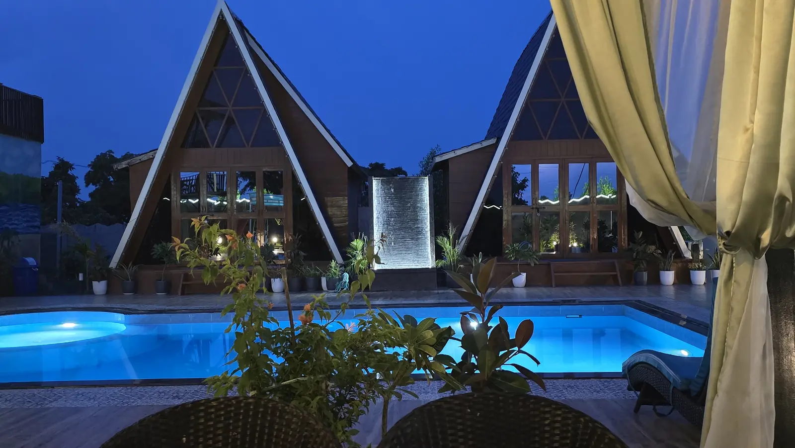 Two A-frame cottages and the lit water wall across the pool at blue hour, framed by a curtain